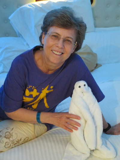 Photo shows Dona sitting on her bed and smiling, pointing to a small white terry-cloth penguin.